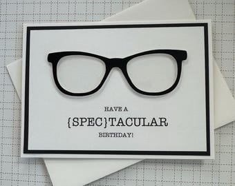 May include: A white card with a black border and a pair of black glasses. The text on the card reads "Have a [SPEC]TACULAR BIRTHDAY!"