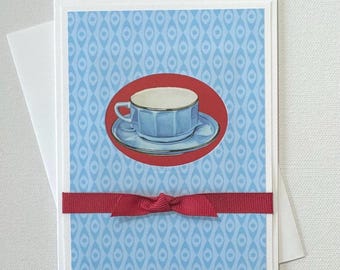 May include: Greeting card featuring a blue teacup and saucer illustration inside a red oval, set against a blue patterned background. A red ribbon is tied across the card. Includes a white envelope.