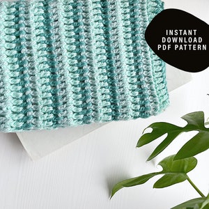 May include: A crochet blanket with a gradient of light blue and teal colors. The blanket is lying on a white surface with a green plant in the background. The text "INSTANT DOWNLOAD PDF PATTERN" is on a black circle.