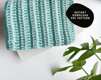 May include: A crochet blanket with a gradient of light blue and teal colors. The blanket is lying on a white surface with a green plant in the background. The text "INSTANT DOWNLOAD PDF PATTERN" is on a black circle.