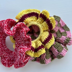 May include: Three crocheted hair scrunchies in different colours and patterns. One is yellow with maroon stripes, one is pink with green stripes, and one is red with white.