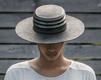May include: A gray fedora-style hat with a wide brim and a layered crown. The hat is worn by a person wearing a light gray t-shirt. The background is a dark gray wooden wall.