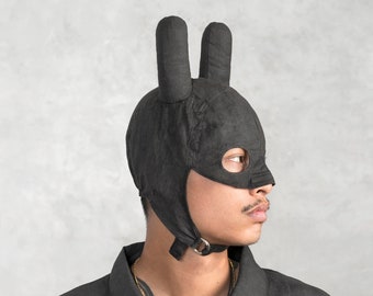 May include: A black fabric mask with bunny ears. The mask covers the eyes and nose and has a small opening for the mouth. The mask is worn by a person with a black shirt.