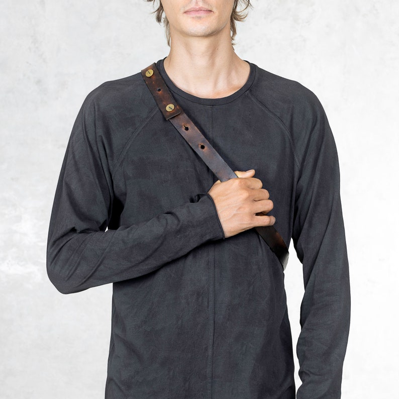 Puede incluir: Un arn&eacute;s de hombro de cuero marr&oacute;n oscuro con remaches color lat&oacute;n. El arn&eacute;s se lleva sobre una camiseta de manga larga gris oscuro. El cuero parece desgastado y la camisa tiene un aspecto texturizado. El estilo general es minimalista y atrevido.