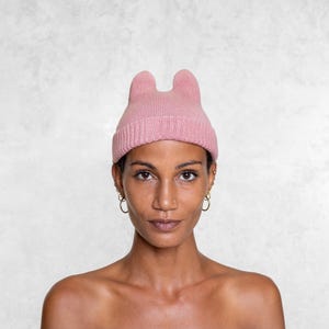 May include: A light pink beanie hat with two upright ear-like protrusions. The hat is made of a ribbed knit material. The person in the image is wearing gold hoop earrings and a nose ring.