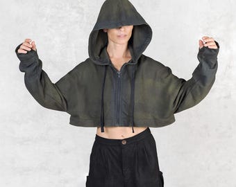May include: A cropped, olive green hooded sweatshirt with a front zipper and long sleeves. The sleeves extend past the hands. The model is also wearing black pants.