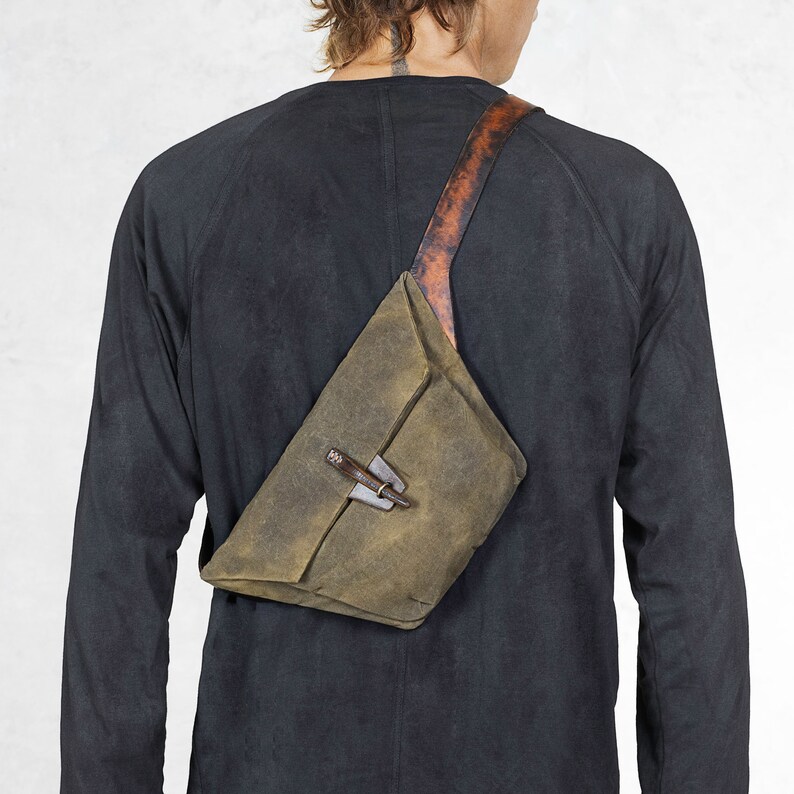 Puede incluir: Bolso bandolera verde oliva con correa de cuero y cierre de metal. El bolso se lleva en diagonal sobre el cuerpo. La correa es marr&oacute;n con un patr&oacute;n texturizado. El bolso tiene forma triangular.