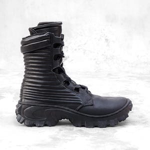 May include: Black combat boot with a ridged shaft and a thick, lugged sole. The boot features a lace-up closure and multiple straps. The upper is made of a matte material, and the boot has a high-top design.