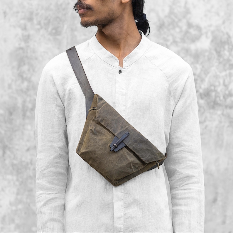 Puede incluir: Bolso bandolera de lona encerada verde oliva con correa gris oscuro y cierre met&aacute;lico. El bolso, de dise&ntilde;o angular, se lleva cruzado, mostrando su est&eacute;tica minimalista.