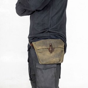 Puede incluir: Ri&ntilde;onera de lona encerada verde oliva con cierre de cuero. La ri&ntilde;onera, de forma rectangular, se lleva en la cintura y tiene una solapa. Est&aacute; sujeta a un cintur&oacute;n de cuero marr&oacute;n. La persona lleva una camisa negra de manga larga y pantalones grises.