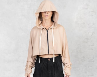 Könnte beinhalten: Ein gecroppter beigefarbener Kapuzenpullover mit durchgehendem schwarzem Reißverschluss und weißen Kordeln. Der Hoodie hat eine große Kapuze und lange Ärmel. Das Model trägt eine schwarze Hose mit hohem Bund. Der Hintergrund ist eine hellgraue Wand.