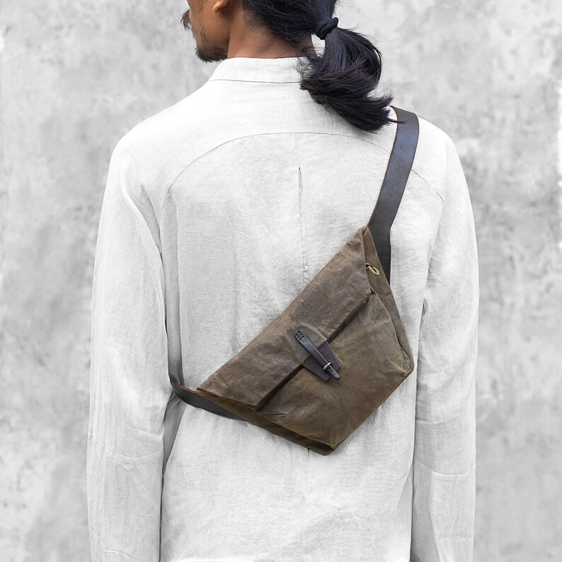 Puede incluir: Un bolso bandolera verde oliva con una correa y detalles de cuero marr&oacute;n oscuro. El bolso tiene forma triangular con cierre de solapa y hebilla. El bolso se lleva sobre el hombro.