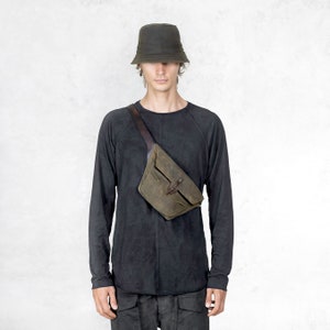 Puede incluir: Una persona con un sombrero de pescador verde oscuro, una camiseta negra de manga larga y una ri&ntilde;onera verde-marr&oacute;n con correa y hebilla de cuero. La ri&ntilde;onera se lleva cruzada sobre el pecho.