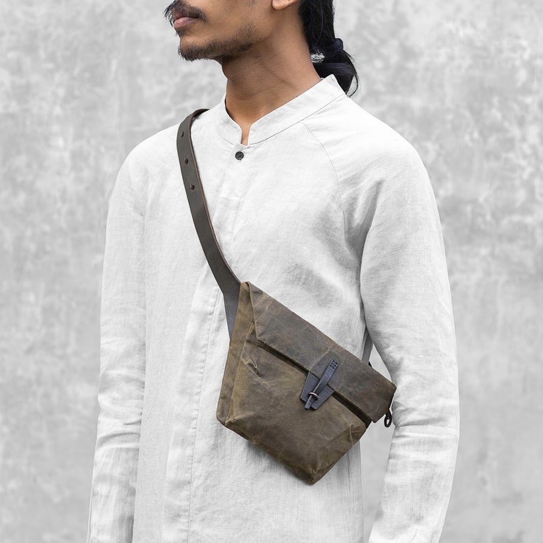 Puede incluir: Bolso bandolera de lona encerada verde oliva con correa de cuero marr&oacute;n oscuro y cierre de hebilla negro. El bolso se lleva sobre el hombro. El bolso tiene forma triangular.