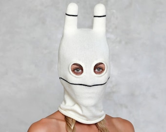 May include: White knitted balaclava with black eye holes and a black line for a mouth. The balaclava has two long bunny ear-like flaps on top.