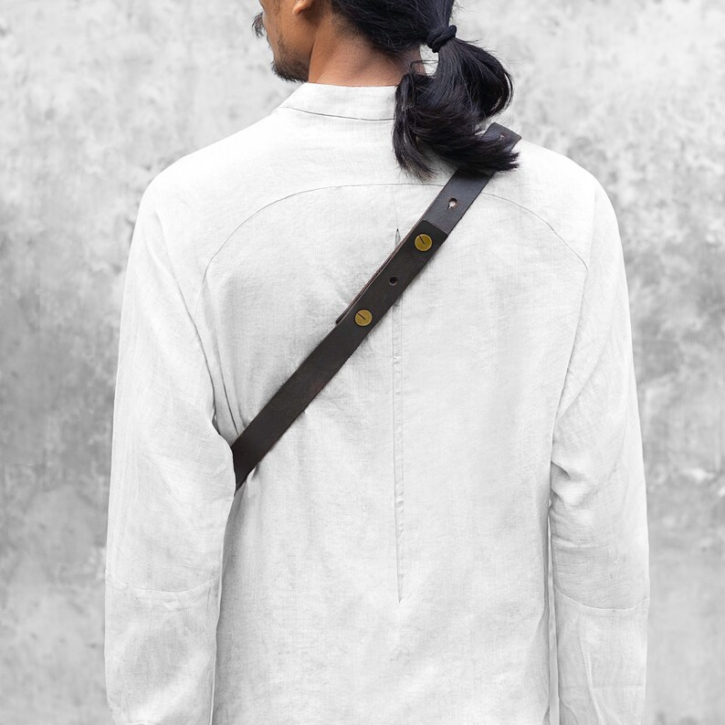 Puede incluir: Una correa de hombro de cuero marr&oacute;n oscuro con remaches color lat&oacute;n se lleva sobre una camisa de lino blanca. La correa est&aacute; en &aacute;ngulo sobre la espalda, lo que sugiere una bolsa o un accesorio. El fondo es una pared gris texturizada.