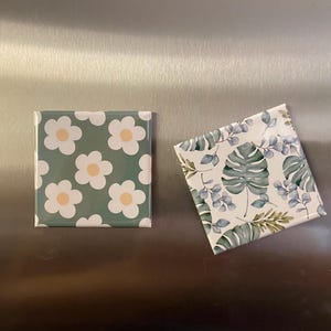 May include: Two square ceramic coasters. One features a pattern of white daisies with yellow centers on a green background. The other has a botanical design with green leaves and blue accents on a white background.