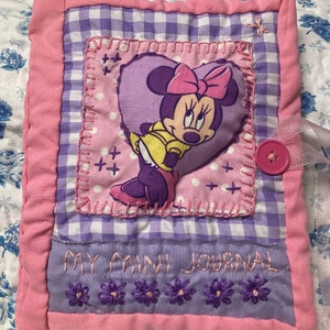 Peut inclure: Un journal matelassé rose et violet avec un motif de Minnie Mouse. Le journal a un bouton rose et les mots "My Mini Journal" brodés sur le devant.