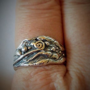 May include: A silver ring with a textured design featuring a swirl and small beads. The ring is worn on a finger.