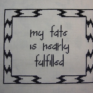 Puede incluir: Obra de arte de punto de cruz sobre tela blanca. El texto "my fate is nearly fulfilled" está centrado dentro de un borde geométrico negro. La rotulación es de un estilo sencillo y manuscrito, y el borde tiene un patrón de zigzag repetitivo.