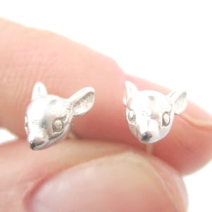 May include: A pair of silver-colored stud earrings shaped like deer heads. The earrings are small and detailed, with visible ears, eyes, and noses. The earrings are held in a person's hand against a white background.