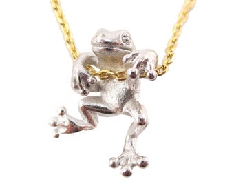 May include: A silver frog pendant with a gold chain necklace. The frog is in a playful pose, with its arms seemingly holding onto the chain. The necklace is set against a white background.