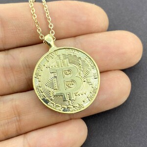 Könnte beinhalten: Goldfarbene Bitcoin-Anhänger-Halskette. Der Anhänger ist rund mit einem Bitcoin-Symbol in der Mitte und aufwendigen Details an den Rändern. Die Kette ist zierlich und ebenfalls goldfarben.
