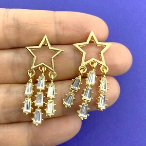 May include: A pair of gold-coloured star earrings. Each earring has an open star design at the top, with three strands of clear, rectangular crystals hanging below. The earrings are set against a blue background.