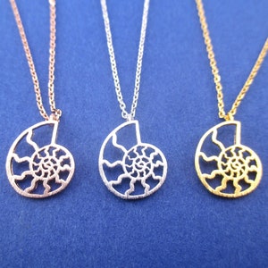 Puede incluir: Tres collares con colgantes de concha marina en oro rosa, plata y oro. Cada colgante cuelga de una fina cadena. Fondo azul.