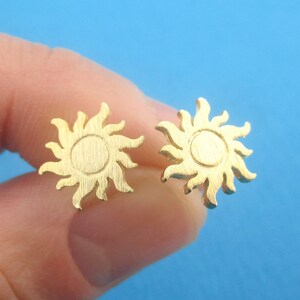 May include: A pair of gold-colored stud earrings. Each earring features a sun design with a circular center and radiating rays. The earrings are held against a blue background.