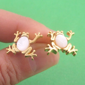May include: A pair of gold-colored frog-shaped earrings. Each earring features a white oval-shaped stone set in a gold-tone metal frame. The frogs have outstretched arms and legs, and are displayed against a green background.
