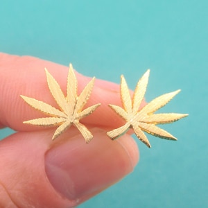 Puede incluir: Un par de pendientes dorados con forma de hojas de cannabis. Los pendientes tienen una superficie texturizada y se muestran sobre un fondo azul claro. Los pendientes son pequeños y delicados.