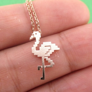 May include: A gold-colored necklace featuring a pixelated flamingo pendant. The pendant is a stylized representation of the bird, with a delicate chain. The necklace is shown against a green background.