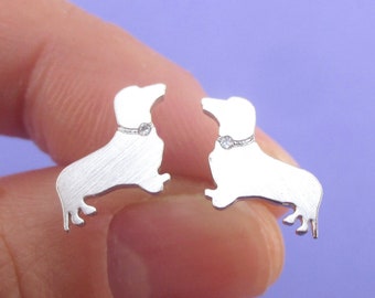 Op de afbeelding: Een paar zilverkleurige teckel oorbellen. Elke oorbel toont een silhouet van een teckel met een kleine, ronde, heldere edelsteen op de halsband. Op een paarse achtergrond.