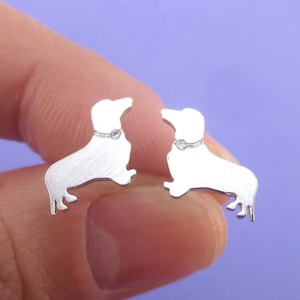 May include: A pair of silver-colored dachshund dog earrings. Each earring features a silhouette of a dachshund with a small, round, clear gem on its collar. The earrings are displayed against a purple background.