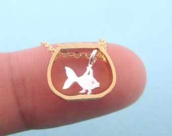 May include: A gold-colored necklace pendant shaped like a fishbowl, with a silver-colored fish inside. The pendant is suspended from a delicate gold chain. The background is a light blue.