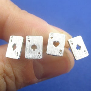 Könnte beinhalten: Vier silberfarbene Spielkarten-Anhänger, jeweils mit einem Ass-Design. Die Farben sind Karo, Pik, Herz und Kreuz. Die rechteckigen Anhänger haben eine gebürstete Metalloberfläche, vor blauem Hintergrund.
