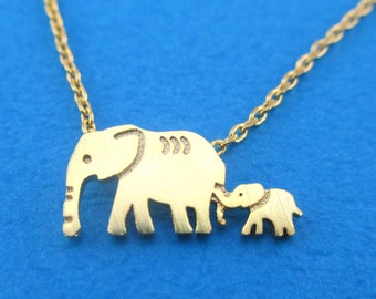 May include: A gold-colored necklace featuring a pendant with two elephant silhouettes, a larger one and a smaller one, connected by a delicate chain. The elephants are side-by-side, with the smaller one next to the larger one. The background is a solid blue.