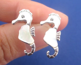 May include: A pair of silver seahorse earrings. Each earring features a seahorse design with a white, iridescent body and a detailed silver head and tail. The earrings are held against a blue background.
