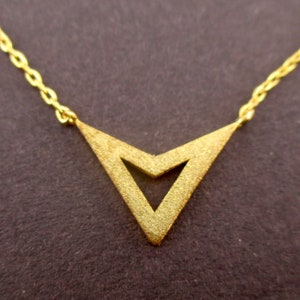 May include: A gold necklace featuring a V-shaped pendant. The pendant has a textured finish and a central cutout. The chain is fine and gold. The necklace is set against a dark brown backdrop.