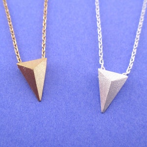 May include: Two necklaces with geometric triangle pendants. One pendant is gold-coloured, the other is silver-coloured. Both pendants hang from delicate chain necklaces. The background is a solid blue.