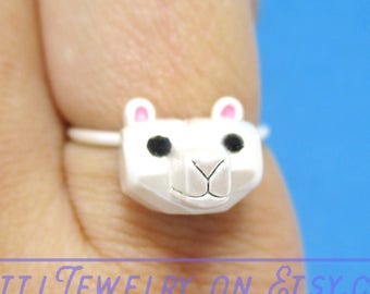 May include: A white llama-shaped ring with black eyes and pink inner ears. The ring is on a finger, with the text "Atti Jewelry on Etsy.com" in the background. The llama has a geometric design.