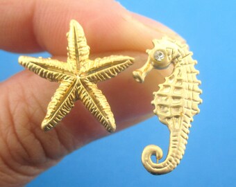 May include: A pair of gold-colored stud earrings. One earring is a starfish, and the other is a seahorse with a small, clear gem. The earrings are likely made of metal and are designed to be worn in pierced ears.