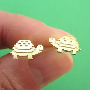 May include: A pair of gold-colored turtle stud earrings. Each earring features a pixelated design, giving them a retro video game aesthetic. The earrings are held against a green background.