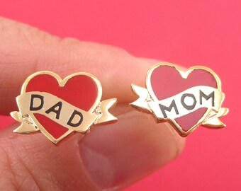 Könnte beinhalten: Ein Paar goldfarbene herzförmige Ohrringe. Jeder Ohrring hat ein rotes Herz mit einem Banner. Auf einem Ohrring steht "DAD" und auf dem anderen "MOM" in schwarzer Schrift. Die Ohrringe sind vor rotem Hintergrund.