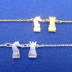 Puede incluir: Dos delicados collares, uno dorado y otro plateado, cada uno con dijes de piezas de ajedrez. El collar dorado muestra un caballo y una torre, mientras que el collar plateado también presenta un caballo y una torre. El fondo es azul liso.