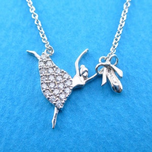 May include: A silver-colored necklace featuring a ballerina pendant with a sparkling, gem-studded dress. The pendant includes a ballet slipper charm. The necklace is displayed against a blue background.