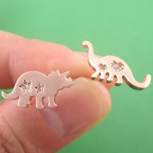 May include: Rose gold dinosaur stud earrings. One earring is a Triceratops with star-shaped cutouts and the other is a Brontosaurus with heart-shaped cutouts. Both earrings have small, clear gemstones.