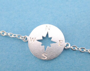 May include: A silver-colored necklace featuring a compass design. The compass has a star-shaped cutout in the center and the cardinal directions are marked. The necklace chain is delicate and the background is a light blue.
