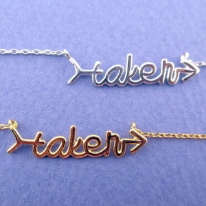 Könnte beinhalten: Zwei Halsketten mit dem Wort "taken" in Schreibschrift, jeweils mit einem Pfeildesign. Eine Halskette ist silberfarben, die andere goldfarben. Beide Halsketten befinden sich auf einer zarten Kette vor einem lila Hintergrund.